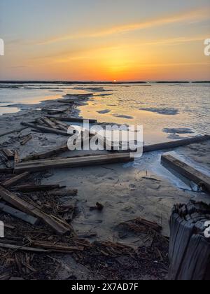 Ein wunderschöner Sonnenuntergang über einem ruhigen Strand mit verstreutem Treibholz, ruhigem Wasser und atemberaubenden Reflexionen, die eine friedliche und ruhige Atmosphäre schaffen. Stockfoto