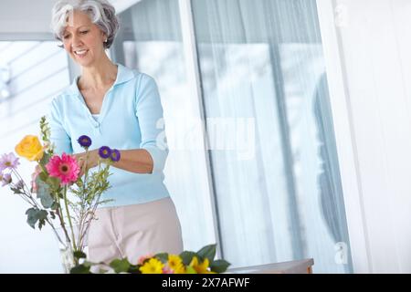 Reife Frau, Vase und Blumen zu Hause mit Gedanken, Lächeln und natürlicher Blumendekoration am Morgen. Senior Person, glücklich und stilvoll mit Blumenstrauß Stockfoto