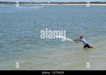 Ein Fischer mit einem zerrissenen Strohhut wirft sein Netz in die Gewässer von Ponce Inlet, Florida, mit Booten und einem felsigen Steg im Hintergrund. Stockfoto