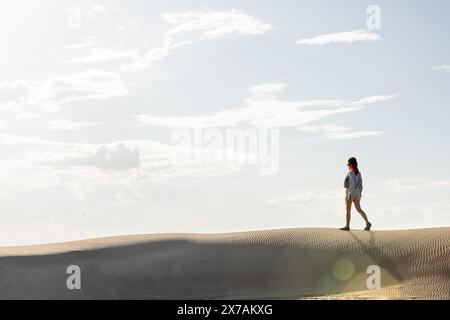 Frau in der Wüste, die bei Sonnenuntergang durch Dünen geht, mit der Sonne gegen den Himmel, kopiert Raum am Himmel Stockfoto