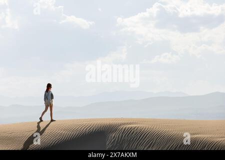 Frau in der Wüste, die bei Sonnenuntergang durch Dünen geht, mit der Sonne gegen den Himmel, kopiert Raum am Himmel Stockfoto