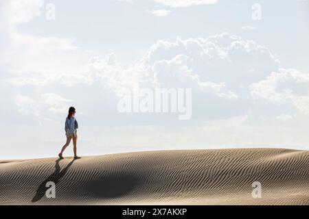 Frau in der Wüste, die bei Sonnenuntergang durch Dünen geht, mit der Sonne gegen den Himmel, kopiert Raum am Himmel Stockfoto