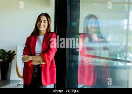 Die junge Frau steht selbstbewusst mit einem Lächeln in einem stilvollen roten Blazer und lehnt sich in einem modernen Bürobereich an ein Glasfenster Stockfoto
