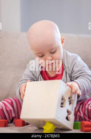 Baby mit Krebs und Haarausfall durch Chemotherapie, das mit Holzspielzeug auf dem Sofa spielt Stockfoto