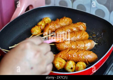 Asiatische Frauenhände verteilen Grillsoße auf Würstchen und Fleischbällchen in einer Grillpfanne Stockfoto