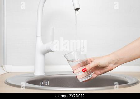 Frau gießt Wasser in ein Glas in der Küche. Der Mensch gießt Wasser aus einem Wasserhahn in ein Glas. Transparentes Glas Wasser. Glas mit sauberem Wasser Stockfoto
