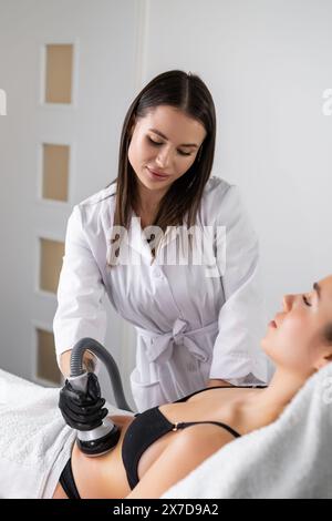 Lächelnde Kundin kommt in den Salon, um einen geformten Magen zu bekommen. Lady fühlt sich gut an, wenn sie Vakuumkavitation durchläuft, die von einem qualifizierten Spezialisten durchgeführt wird Stockfoto