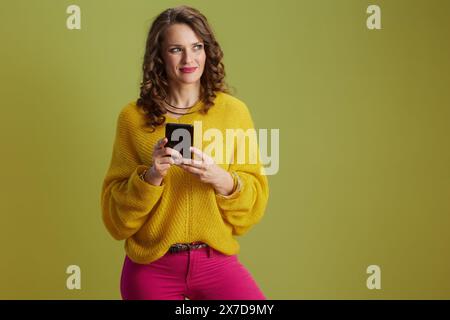 Nachdenklich geschnittene Frau auf grünem Hintergrund in gelbem Pullover mit Smartphone. Stockfoto