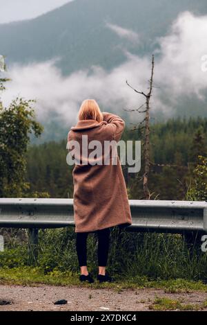 Die schöne Fotografin fotografiert norwegische Berge mit Nebel an einem regnerischen Herbsttag am Straßenrand durch einen Metallzaun. Stockfoto