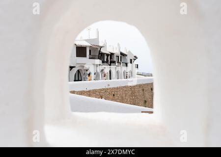 Eine typische Straßenszene in Binibeca, Menorca, mit Blick durch ein kleines Fenster eines weiß getünchten Hauses. Die bezaubernde mediterrane Architektur mit Menschen Stockfoto
