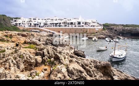 Blick auf Binibeca auf Menorca, Spanien mit seinem Hafen und kleinen Strand mit Felsen im Vordergrund und verankerten Booten Stockfoto