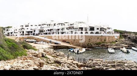 Panoramablick auf Binibeca, Menorca, mit bezauberndem Dorf und Hafen. Im Vordergrund, Felsen und vertäute Boote des malerischen Mediterr Stockfoto