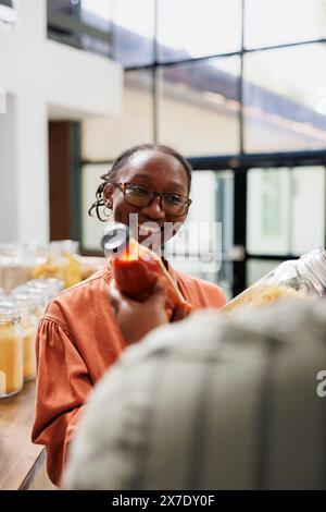 Umweltfreundlicher afroamerikanischer Landwirt verkauft in Flaschen abgefüllte Bio-Produkte auf dem lokalen Markt. Lächelnde schwarze Kundin, die dem Verkäufer über frisch erntete und nachhaltige Optionen im Geschäft zuhört. Stockfoto