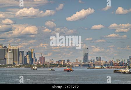 New York, NY, USA - 1. August 2023: Ein Wolkenkratzer am Manhattan Square hinter der Brooklyn Bridge über den East River unter blauer Wolkenlandschaft. Boote weiter Stockfoto