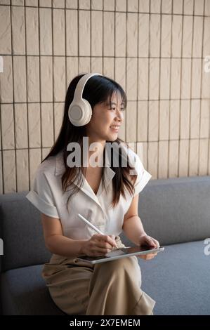 Eine attraktive, sorglose asiatische Frau mit Kopfhörern hört gern Musik und schaut auf die Außenansicht, während sie an ihrem digitalen Tablet i arbeitet Stockfoto