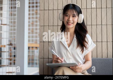 Eine attraktive asiatische Frau oder Geschäftsfrau, die Kopfhörer trägt und Musik hört, während sie in einem Café an ihrem digitalen Tablet arbeitet und in die Kamera lächelt. Stockfoto