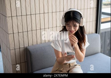 Eine schöne, positive asiatische Frau verwendet ihr Smartphone, liest Nachrichten oder scrollt in einer Musik-App, während sie Musik auf ihrem Kopf hört Stockfoto