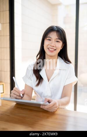 Eine schöne, positive asiatische Geschäftsfrau lächelt in die Kamera, während sie einen Stift und ein digitales Tablet hält und von einem Café aus aus arbeitet Stockfoto