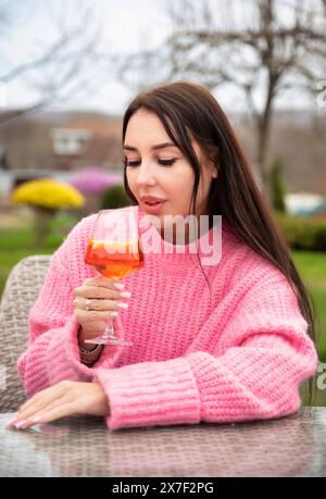 Vertikales Porträt einer jungen russischen Frau, die Cocktail-aperol-Spritz trinkt, draußen im rosa Pullover sitzt Stockfoto