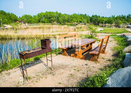 Brazier mit Brennholz mit Holztisch und Bank auf dem Hintergrund des Flusses Stockfoto