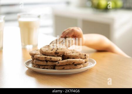 Eine Kinderhand, die nach einem Schokoladenchip-Keks greift, mit einem Glas Milch im Hintergrund Stockfoto