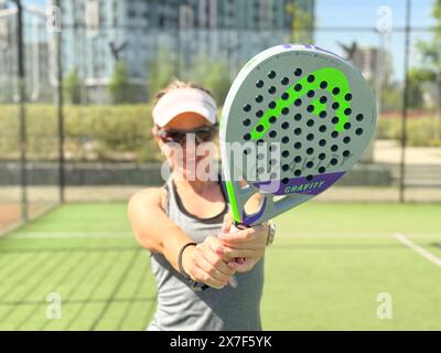 Ukraine Kiew, 03. Mai 2024. Porträt einer lächelnden Frau mit Padel-Tennisschlägerkopf auf dem Hof Stockfoto