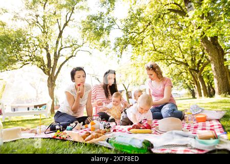 Mütter und Babys genießen ein Picknick im Freien im Park, sitzen auf einer Picknickdecke und bereiten Speisen und Getränke zu. Stockfoto