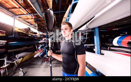 Portrat eines jungen Kanufahrers, der mitten in gestapelten Kanus steht. Das Konzept des Kanufahrens als dynamischer und abenteuerlicher Sport. Stockfoto