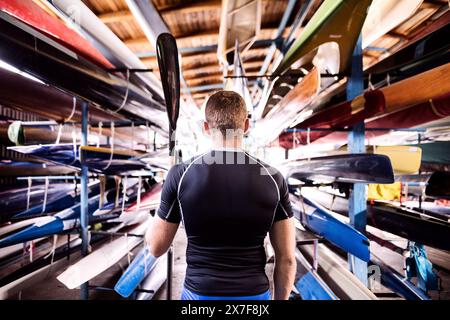 Portrat eines jungen Kanufahrers, der mitten in gestapelten Kanus steht. Das Konzept des Kanufahrens als dynamischer und abenteuerlicher Sport. Stockfoto