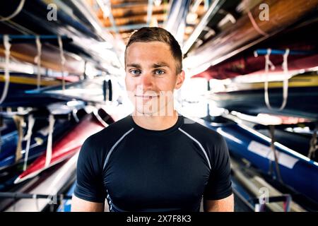 Portrat eines jungen Kanufahrers, der mitten in gestapelten Kanus steht. Das Konzept des Kanufahrens als dynamischer und abenteuerlicher Sport. Stockfoto