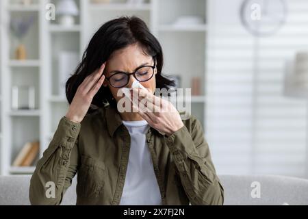 Nahaufnahme einer kranken jungen Frau, die zu Hause auf dem Sofa sitzt, ihren Kopf mit der Hand hält und ihre Nase mit einer Serviette von einer Erkältung abwischt. Stockfoto