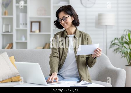 Eine fokussierte junge Frau sitzt bequem auf einer Couch und arbeitet an ihrem Laptop, während sie ein Dokument hält. Sie ist in einem gut beleuchteten Wohnzimmer mit modernem Dekor. Stockfoto