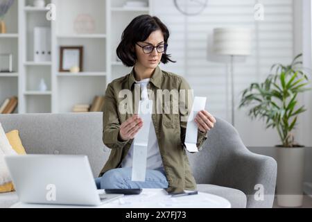 Besorgte und ernste junge Frau sitzt zu Hause auf dem Sofa, überprüft Belege für Einkäufe und Ausgaben, mit Laptop und Rechner. Stockfoto