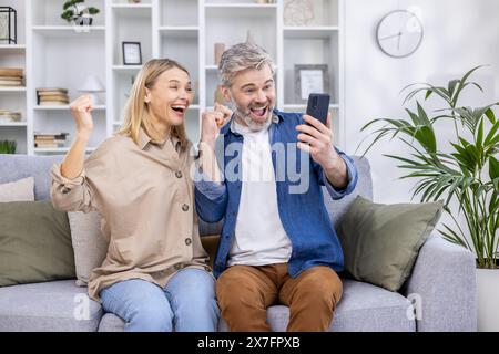 Älteres Paar feiert freudig Erfolg, während es auf das Smartphone blickt, Aufregung und Glück zeigt und auf einer Couch in einem hellen Wohnzimmer sitzt. Stockfoto