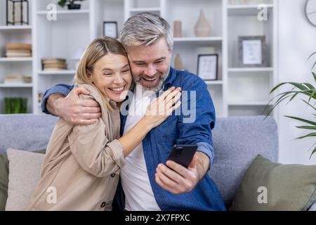 Fröhliches Paar mittleren Alters, das sich umarmt und ein Selfie auf einem Smartphone macht, während es auf der Couch in einem gemütlichen Wohnzimmer sitzt. Stockfoto