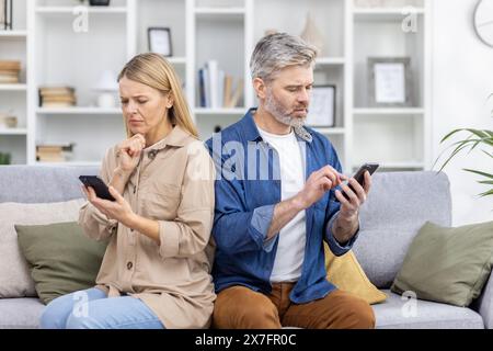 Reife Ehepaare, die mit Smartphones mit ernstem Ausdruck Rücken auf einer Couch sitzen, konzentrierten sich beide auf ihre Geräte in einem modernen Wohnraum. Stockfoto