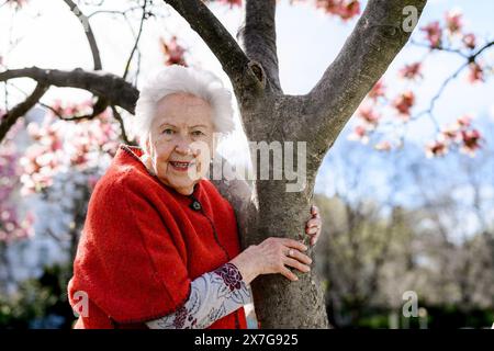 Porträt einer älteren Frau, die am Magnolienbaum steht, im Park, einen entspannenden Moment hat. Großmutter genießt warmes Frühlingswetter. Stockfoto