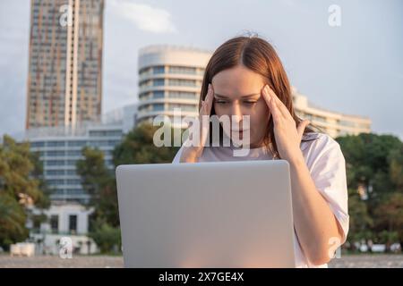 Gestresste Freiberuflerin, die im Freien an einem Laptop arbeitet, mit Kopfschmerzen, sieht müde und überfordert aus. Frustrierte Frauen spüren Schmerzen im Kopf Stockfoto