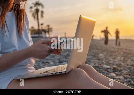 Nahaufnahme einer Frau, die mit Laptop und Handy am Strand am Meer während des Sonnenuntergangs arbeitet. Freiberufliche Frauen arbeiten online mit dem Smartphone und Stockfoto
