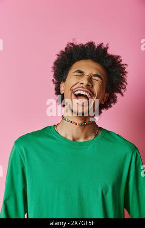 Ein fröhlicher junger Afroamerikaner mit einem lockigen Afro lacht, während er ein grünes Hemd auf rosa Hintergrund trägt. Stockfoto