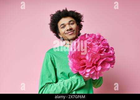 Ein fröhlicher, lockiger Afroamerikaner in Freizeitkleidung hält eine große rosa Blume vor einem rosa Hintergrund. Stockfoto