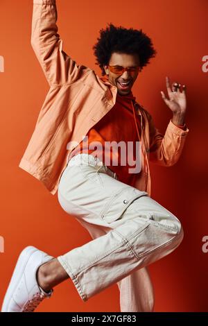 Lockiger afroamerikanischer Mann in trendigem Outfit und Sonnenbrille, der vor orangefarbenem Hintergrund energisch in die Luft springt. Stockfoto