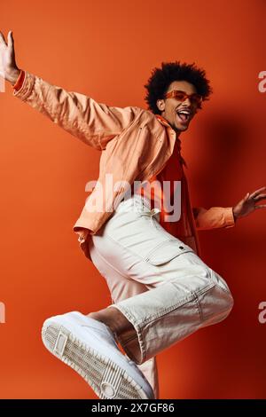 Ein energiegeladener junger afroamerikanischer Mann springt mit ausgestreckten Armen in die Luft und strahlt Freude und Freiheit vor einem orangen Hintergrund aus. Stockfoto