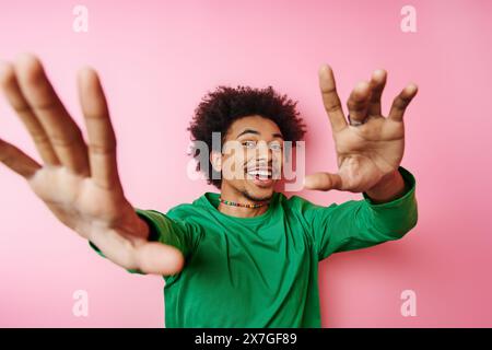 Junger Mann mit lockigen Haaren in lässiger Kleidung, der aufgeregt die Hände auf rosa Hintergrund hebt. Stockfoto