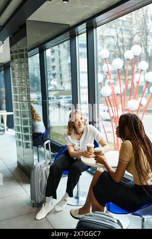 Zwei multikulturelle Kollegen, verschiedene Geschäftsfrauen, die an einem Tisch in einem Hotel sitzen und sich vor einem Fenster intensiv unterhalten. Stockfoto