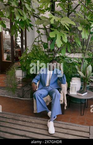 Ein gutaussehender Afroamerikaner im blauen Anzug sitzt auf einer Bank in einem lebhaften grünen Garten. Stockfoto