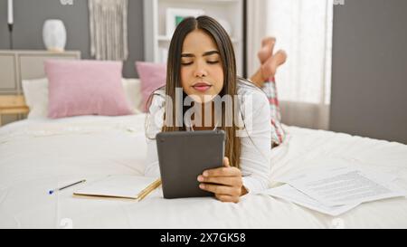 Junge hispanische Frau, die auf dem Bett mit Dokumenten und Tablet in einer gemütlichen Schlafzimmerumgebung liegt Stockfoto