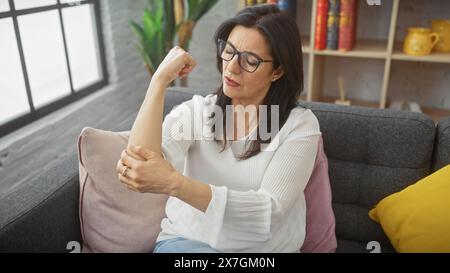 Reife hispanische Frau hat Schmerzen im Ellenbogen, während sie nachdenklich in einem gemütlichen Wohnzimmer sitzt. Stockfoto