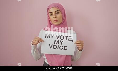 Eine selbstbewusste junge Frau in einem rosafarbenen Hijab hält ein Schild mit „meine Stimme, meine Stimme“ vor einem isolierten rosafarbenen Hintergrund. Stockfoto