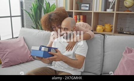 Interrassisches Paar genießt einen gemütlichen Moment in einem modernen Wohnzimmer und teilt ein Überraschungsgeschenk Stockfoto